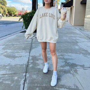 以下が含まれることがあります： 女性は、薄いベージュのトレーナーを着ています。トレーナーには、茶色の文字で「LAKE LIFE」と書かれています。彼女は白いスニーカーを履いており、コーヒーカップを持っています。
