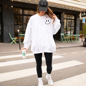 May include: A white sweatshirt with a black smiley face graphic and the text "Good Vibes Only". The person is wearing black leggings and a black baseball cap with white text.