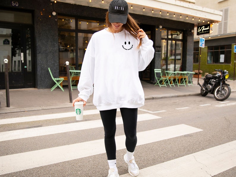 May include: A white sweatshirt with a black smiley face graphic and the text "Good Vibes Only". The person is wearing black leggings and a black baseball cap with white text.