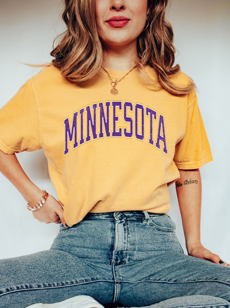 Peut inclure: Une femme portant un t-shirt jaune avec le mot "MINNESOTA" imprim&eacute; en lettres violettes et dor&eacute;es. Elle porte &eacute;galement un jean bleu.
