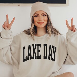 K&ouml;nnte beinhalten: Ein beiger Pullover mit dem Aufdruck "LAKE DAY" in Schwarz auf der Vorderseite. Das Model tr&auml;gt eine wei&szlig;e M&uuml;tze und zeigt mit beiden H&auml;nden das Peace-Zeichen.