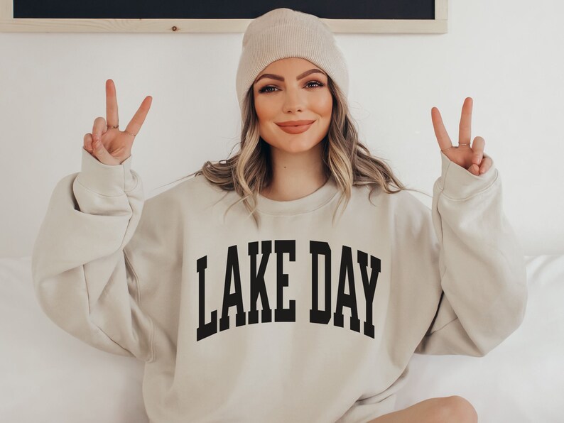 K&ouml;nnte beinhalten: Ein beiger Pullover mit dem Aufdruck "LAKE DAY" in Schwarz auf der Vorderseite. Das Model tr&auml;gt eine wei&szlig;e M&uuml;tze und zeigt mit beiden H&auml;nden das Peace-Zeichen.