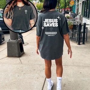 May include: A dark gray oversized t-shirt with the text "JESUS SAVES" and a cross printed on the back. The shirt has a distressed look and features a white scripture quote below the main text. The scripture quote is "For God so loved the world that he gave his only begotten Son, that whosoever believeth in him should not perish but have everlasting life. John 3:16-17."