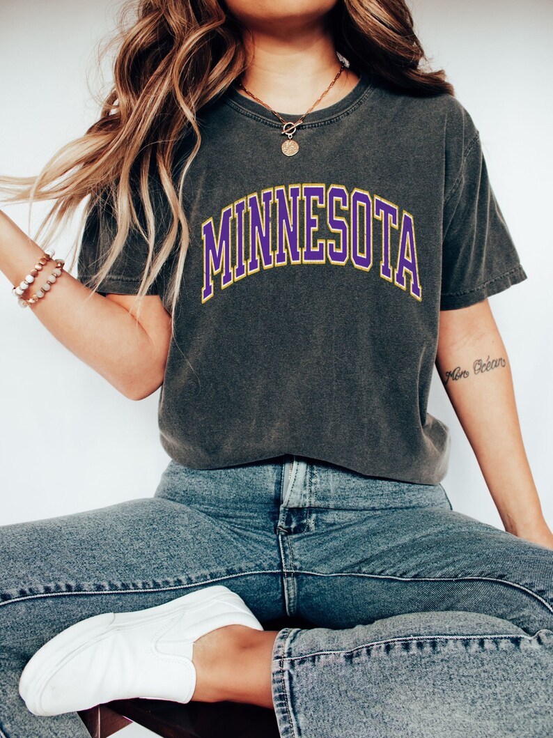 Peut inclure: Un t-shirt gris anthracite avec un graphisme "MINNESOTA" en violet et jaune.