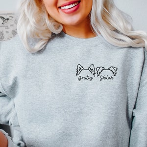 K&ouml;nnte beinhalten: Grauer Pullover mit einer schwarzen Linienzeichnung von zwei Hundefiguren und den Namen "Brutus" und "Shiloh" darunter.