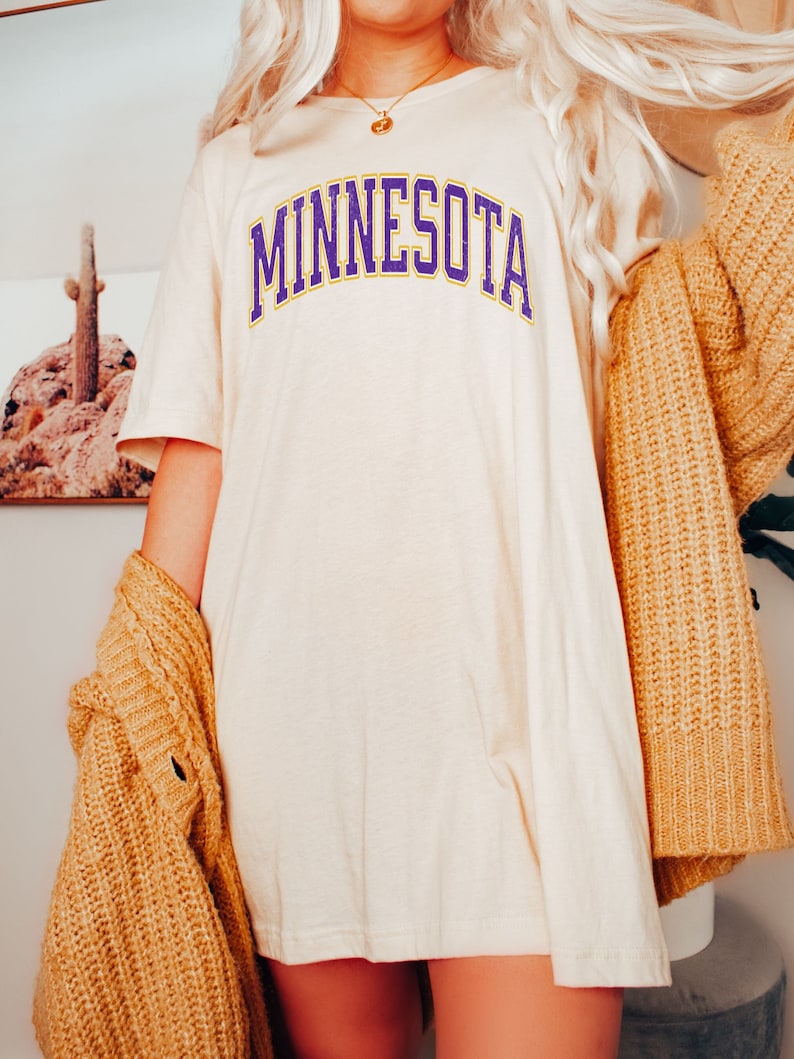 Peut inclure: Un t-shirt blanc avec un imprim&eacute; "MINNESOTA" en violet et jaune. Le t-shirt est port&eacute; avec un cardigan marron.