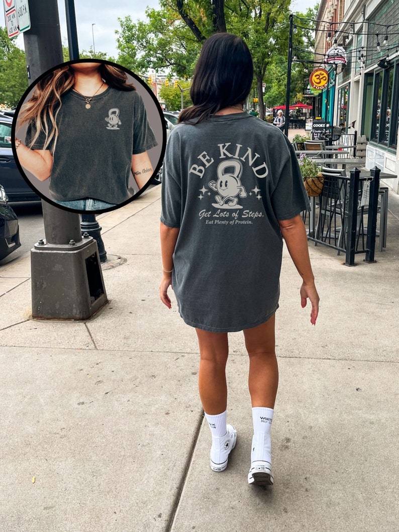 Peut inclure: Un t-shirt gris anthracite avec un graphique d'un halt&egrave;re de kettlebell de dessin anim&eacute; avec le texte "Be Kind, Get Lots of Steps, Eat Plenty of Protein." au dos.