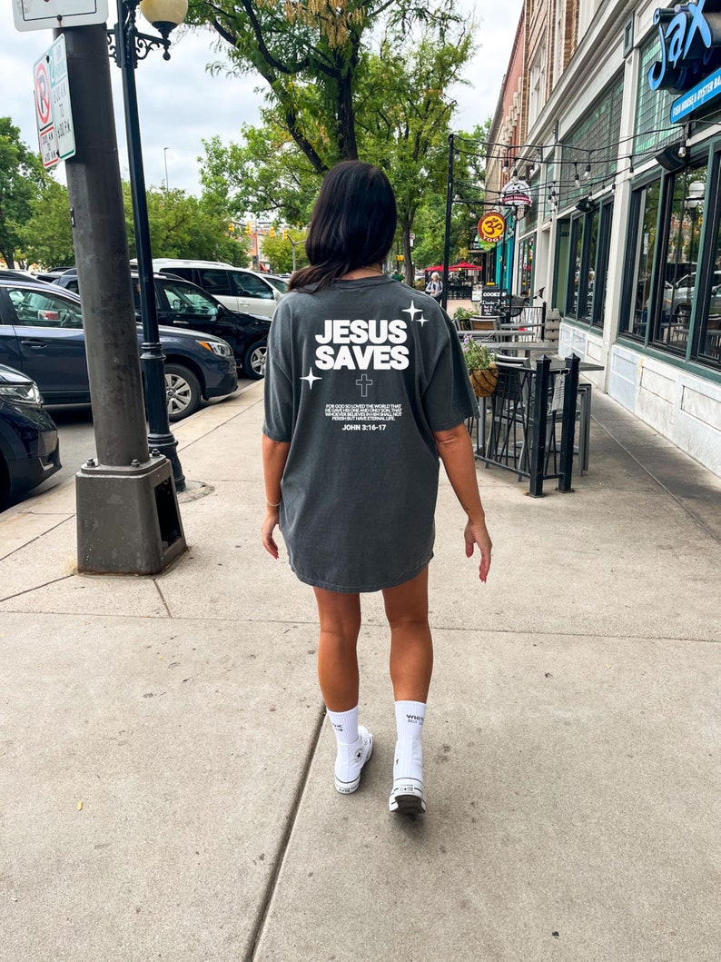 May include: A person wearing a dark gray t-shirt with the text "JESUS SAVES" and a white cross. The shirt also includes the text "For God so loved the world that he gave his one and only Son, that whoever believes in him shall not perish but have eternal life." and "John 3:16-17". The person is wearing white socks and white sneakers.