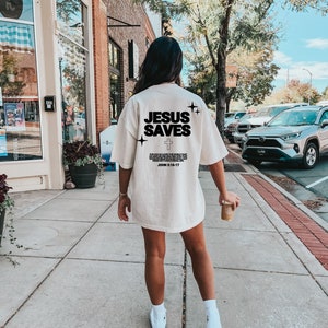 May include: A white t-shirt with the text "JESUS SAVES" and a cross printed on the back. The text is in black and the shirt has a distressed look. The shirt is being worn by a person walking down a sidewalk.