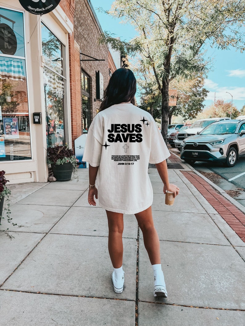 May include: A white t-shirt with the text "JESUS SAVES" and a cross printed on the back. The text is in black and the shirt has a distressed look. The shirt is being worn by a person walking down a sidewalk.