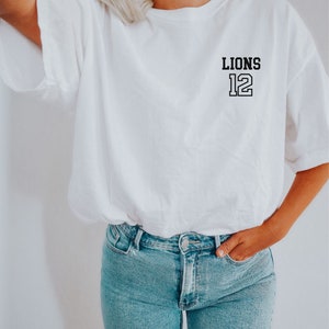 May include: White t-shirt with a black graphic that says "LIONS 12" on the left chest.