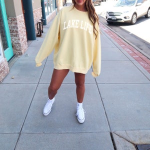May include: A pale yellow sweatshirt with the text "LAKE LIFE" in white letters. The sweatshirt is oversized and worn with white high-top sneakers.