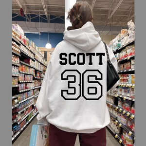 May include: White hoodie with black text that says "SCOTT 36" on the back. The person is wearing the hoodie in a grocery store.