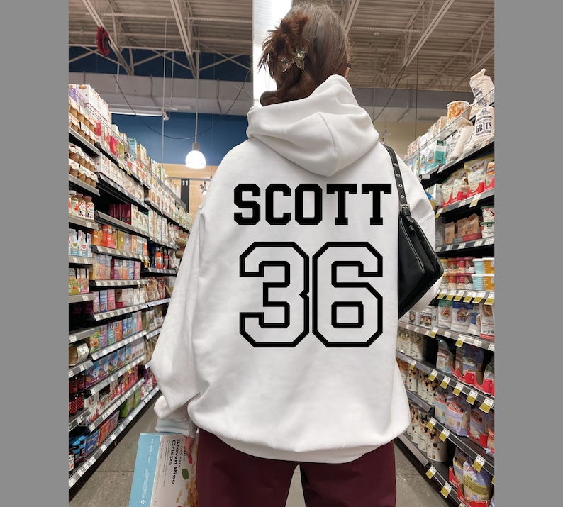 May include: White hoodie with black text that says "SCOTT 36" on the back. The person is wearing the hoodie in a grocery store.