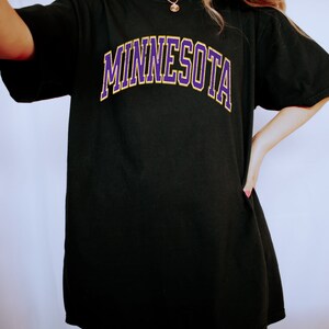 Peut inclure: T-shirt noire surdimensionn&eacute;e avec un graphisme "MINNESOTA" en violet et jaune.