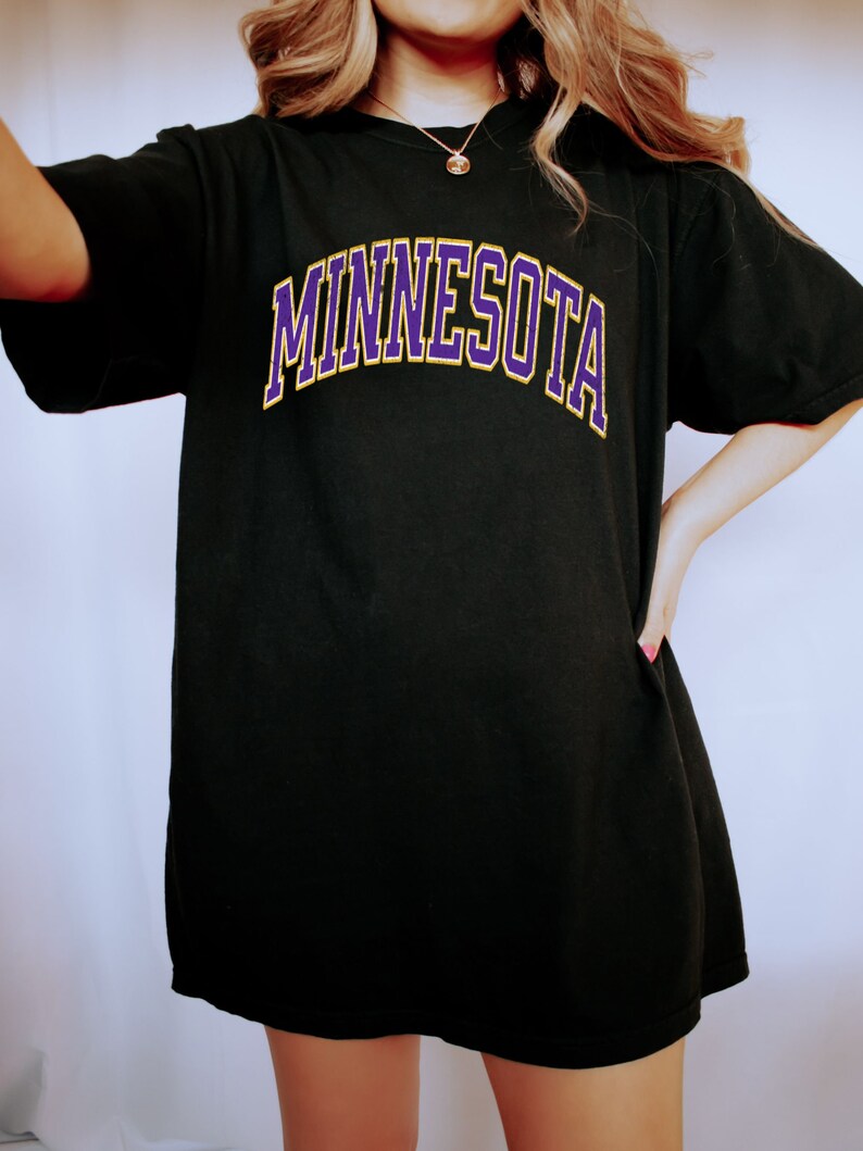Peut inclure: T-shirt noire surdimensionn&eacute;e avec un graphisme "MINNESOTA" en violet et jaune.