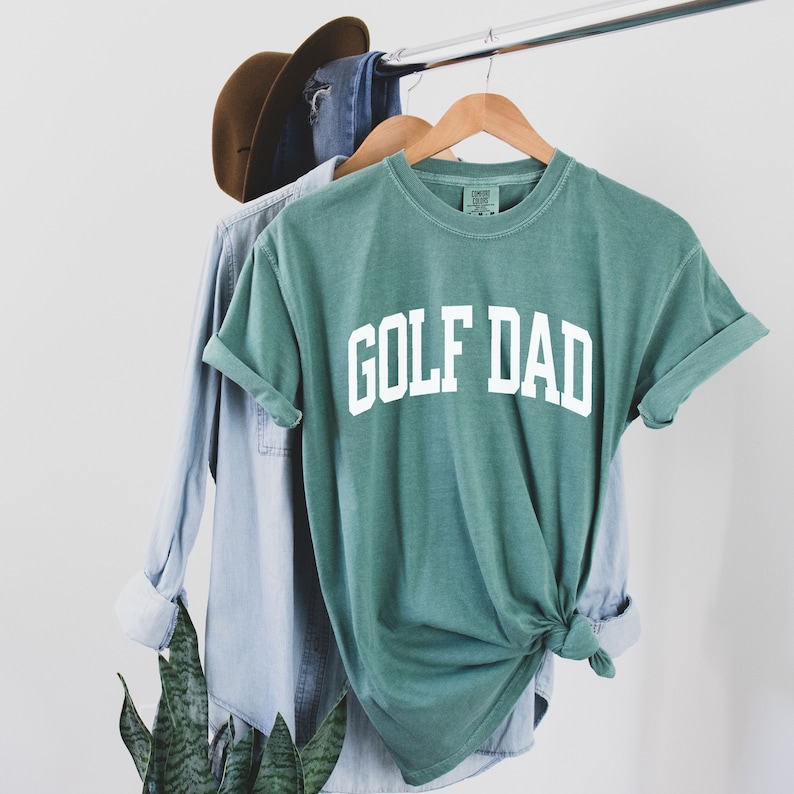 May include: A green t-shirt with the text "GOLF DAD" in white letters. The shirt is hanging on a hanger and is slightly creased.
