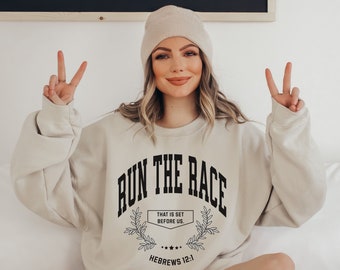 May include: A beige sweatshirt with the text "RUN THE RACE" in black, with a smaller text box below that says "THAT IS SET BEFORE US." and "HEBREWS 12:1".