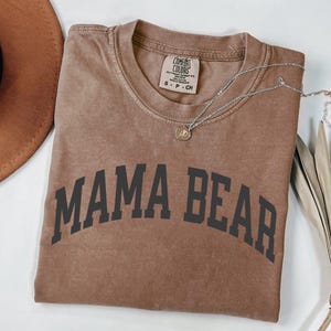 May include: A brown t-shirt with the words "MAMA BEAR" printed in black block letters. The shirt has a crew neck and short sleeves. A silver necklace with a small pendant is draped across the shirt.