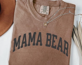 May include: A brown t-shirt with the words "MAMA BEAR" printed in black block letters. The shirt has a crew neck and short sleeves. A silver necklace with a small pendant is draped across the shirt.