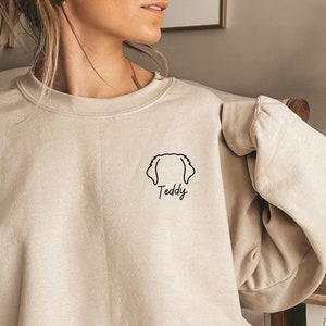 K&ouml;nnte beinhalten: Ein beiger Pullover mit einer schwarzen Umrandung eines Hundekopfes und dem Wort "Teddy" darunter.