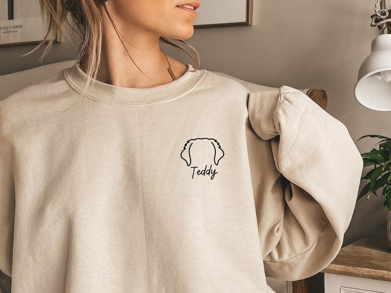 K&ouml;nnte beinhalten: Ein beiger Pullover mit einer schwarzen Umrandung eines Hundekopfes und dem Wort "Teddy" darunter.