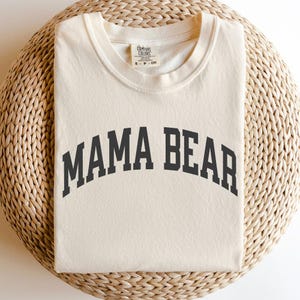 May include: Cream-colored t-shirt with the text "MAMA BEAR" in a dark gray, arched font. The shirt is folded and rests on a woven, light brown surface. The tag reads "COMFORT COLORS".