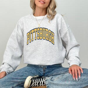 Może przedstawiać: Jasnoszara bluza z napisem "PITTSBURGH" w ż&oacute;łtych literach z czarnym obrysem. Bluza ma długie rękawy i okrągły dekolt.