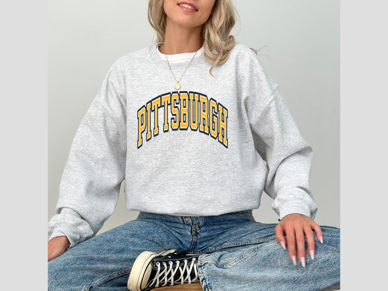 Może przedstawiać: Jasnoszara bluza z napisem "PITTSBURGH" w ż&oacute;łtych literach z czarnym obrysem. Bluza ma długie rękawy i okrągły dekolt.