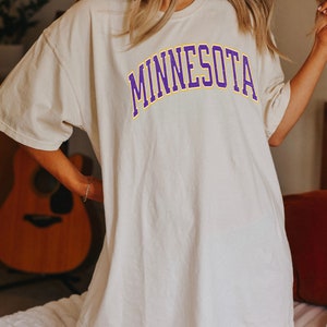 Peut inclure: Un t-shirt blanc surdimensionn&eacute; avec un graphisme "MINNESOTA" en violet et jaune.