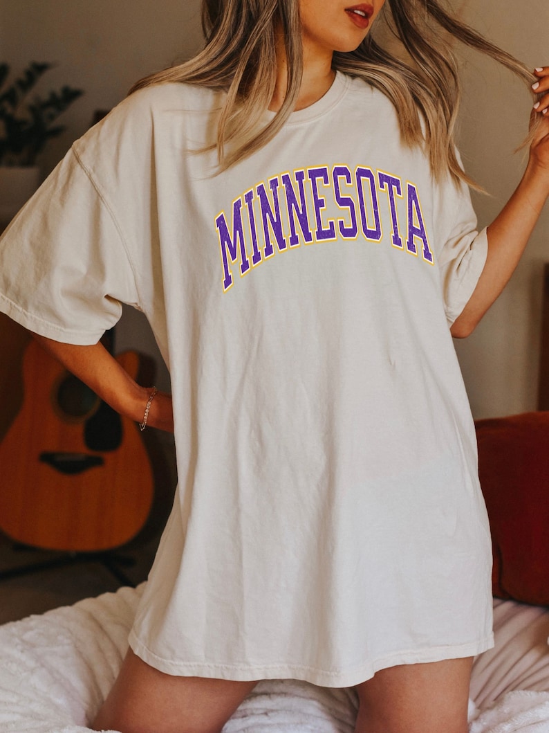 Peut inclure: Un t-shirt blanc surdimensionn&eacute; avec un graphisme "MINNESOTA" en violet et jaune.