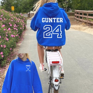 May include: A royal blue hoodie with a white graphic of crossed hockey sticks and the word "FORCE" on the front. The hoodie is also shown on a person wearing it on the back with the name "GUNTER" and the number "24" in white.