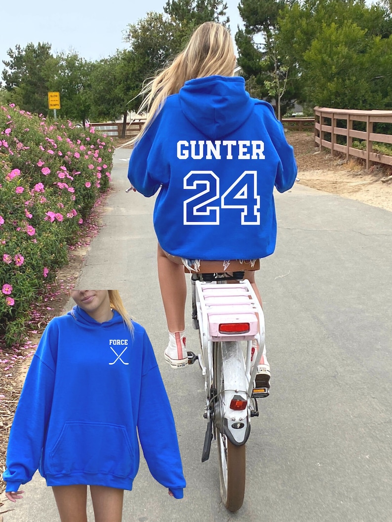 May include: A royal blue hoodie with a white graphic of crossed hockey sticks and the word "FORCE" on the front. The hoodie is also shown on a person wearing it on the back with the name "GUNTER" and the number "24" in white.