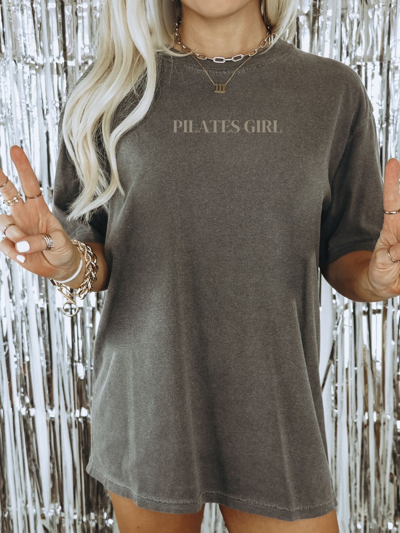 K&ouml;nnte beinhalten: Ein graues Oversize-T-Shirt mit dem Aufdruck "PILATES GIRL" auf der Vorderseite. Das Shirt wird von einer Person mit blonden Haaren und einer goldenen Halskette mit einem r&ouml;mischen Zifferblatt III getragen.