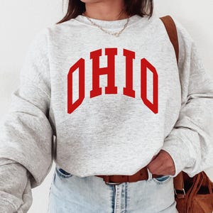 May include: A light grey sweatshirt featuring the word "OHIO" in bold red lettering. The sweatshirt has long sleeves and a relaxed fit. The model is wearing blue jeans and a brown belt.