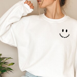 May include: White sweatshirt with a black smiley face and the text "social worker" underneath.