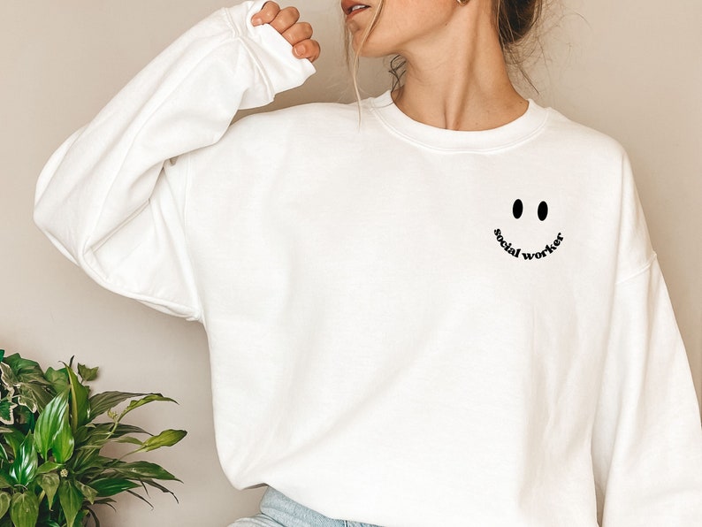 May include: White sweatshirt with a black smiley face and the text "social worker" underneath.