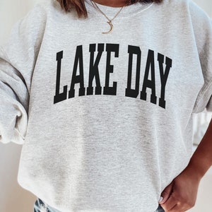 K&ouml;nnte beinhalten: Grauer Pullover mit dem Aufdruck "LAKE DAY" in Schwarz.