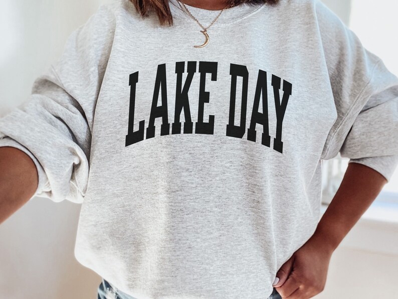 K&ouml;nnte beinhalten: Grauer Pullover mit dem Aufdruck "LAKE DAY" in Schwarz.