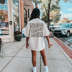 May include: A white t-shirt with the text "Stronger than any disorder" printed on the back in brown letters. The shirt is being worn by a person walking down a sidewalk.
