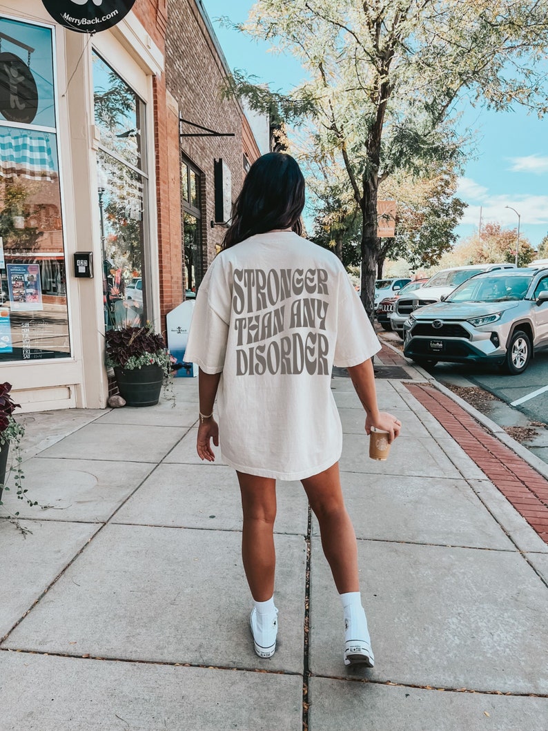 May include: A white t-shirt with the text "Stronger than any disorder" printed on the back in brown letters. The shirt is being worn by a person walking down a sidewalk.