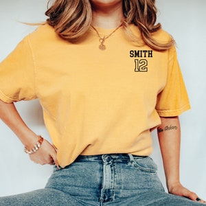 May include: A yellow t-shirt with black text that says "SMITH 12" on the front. The person wearing the shirt is sitting down and wearing blue jeans.