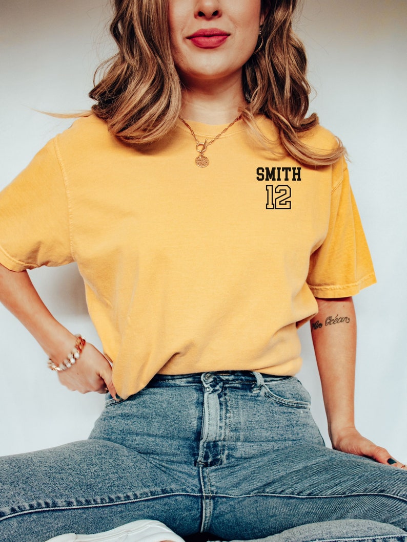 May include: A yellow t-shirt with black text that says "SMITH 12" on the front. The person wearing the shirt is sitting down and wearing blue jeans.