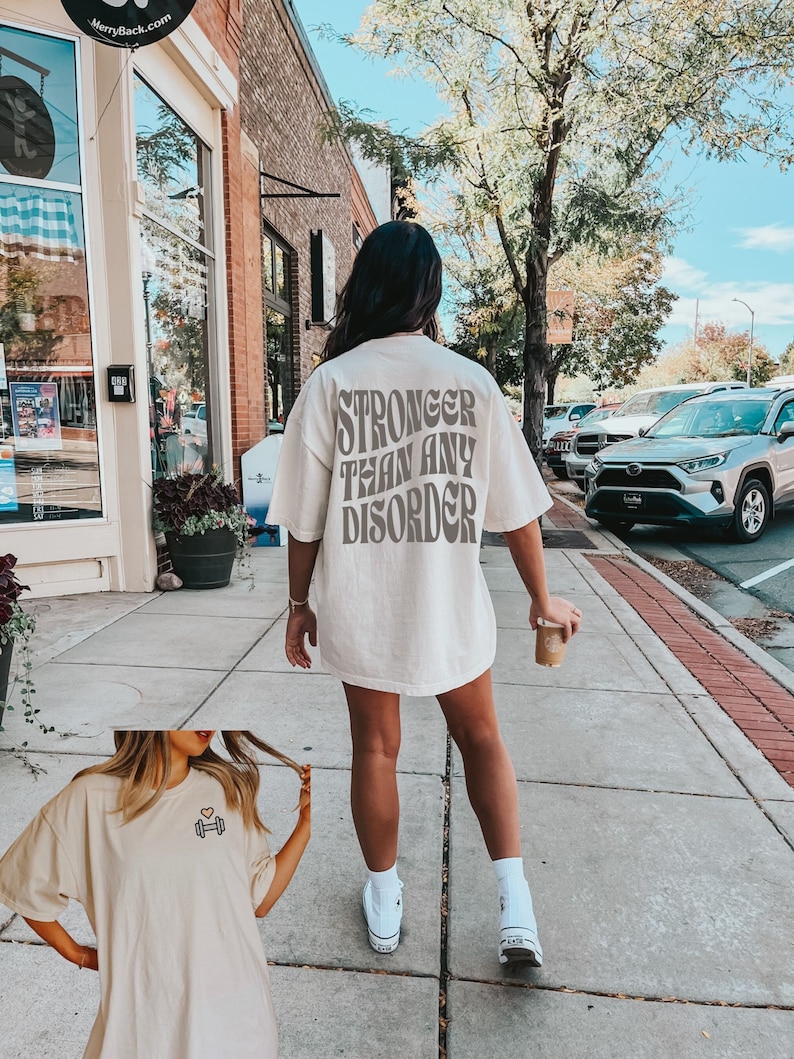 May include: A white oversized t-shirt with the text "Stronger than any disorder" printed on the back. The shirt is being worn by a person walking down a sidewalk. The person is wearing white sneakers.