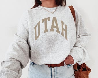 May include: A light grey sweatshirt featuring the word "UTAH" in a tan, arched font. The sweatshirt has long sleeves and a relaxed fit. The person is wearing blue jeans and a brown belt. The image shows a casual style.
