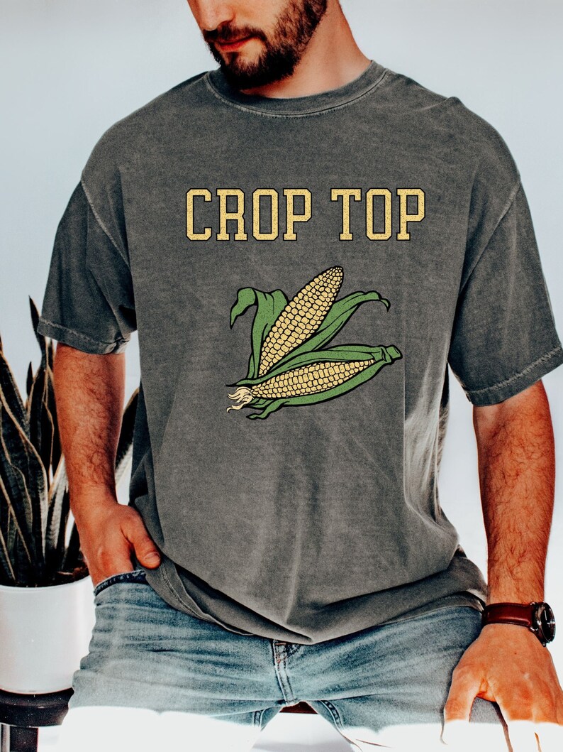 Puede incluir: Una camiseta gris con un aspecto vintage que presenta el texto "CROP TOP" en letras amarillas y un gr&aacute;fico de ma&iacute;z en la mazorca.