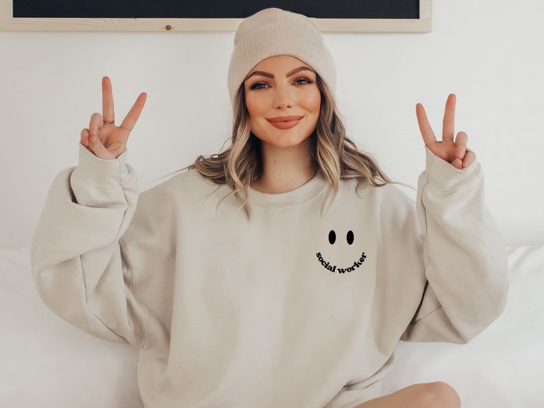 May include: A beige sweatshirt with a black smiley face and the text "social worker" underneath. The person wearing the sweatshirt is smiling and giving a peace sign with both hands.