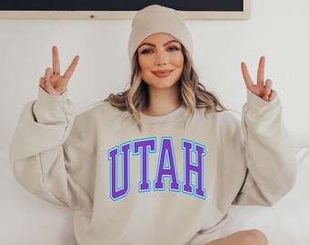 May include: A beige sweatshirt with the word "UTAH" in purple and blue lettering. The person in the image is wearing a beige beanie and making a peace sign with their fingers.