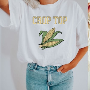 May include: White crop top with the text "CROP TOP" in yellow letters and a graphic of two ears of corn.