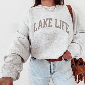 以下が含まれることがあります： アーチ型の大きなフォントで「LAKE LIFE」と書かれたグレーのスウェットシャツ。ブラウンの革のバッグを持った人が着用しています。